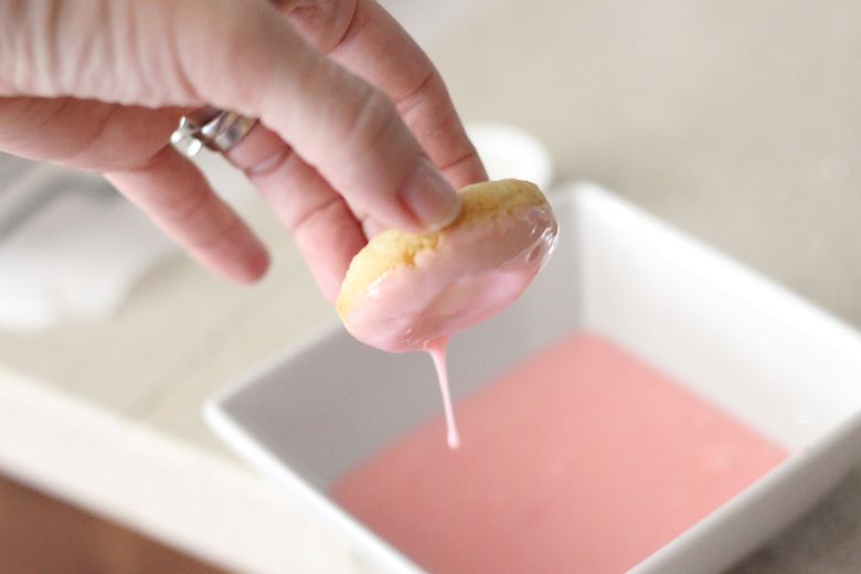 Sugar Cookie Donut held over the bowl of pink almond glaze to allow excess glaze to drip off.