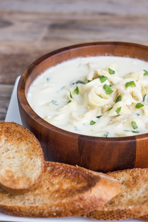 Creamy Spinach And Artichoke Soup With Cheese Tortellini in a bowl that is on a plate with toasted French bread slices.