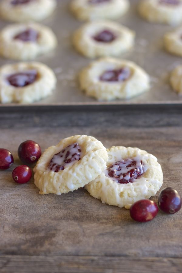 Two Cranberry Thumbprint Cookies With Almond Glaze on a board with cranberries and a baking sheet of the cookies in the background.