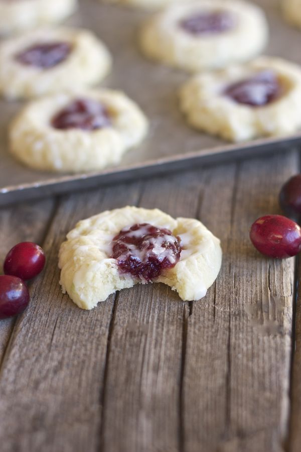 A Cranberry Thumbprint Cookie With Almond Glaze with a bite taken out of it on a board with cranberries and cookies on a baking sheet in the background.