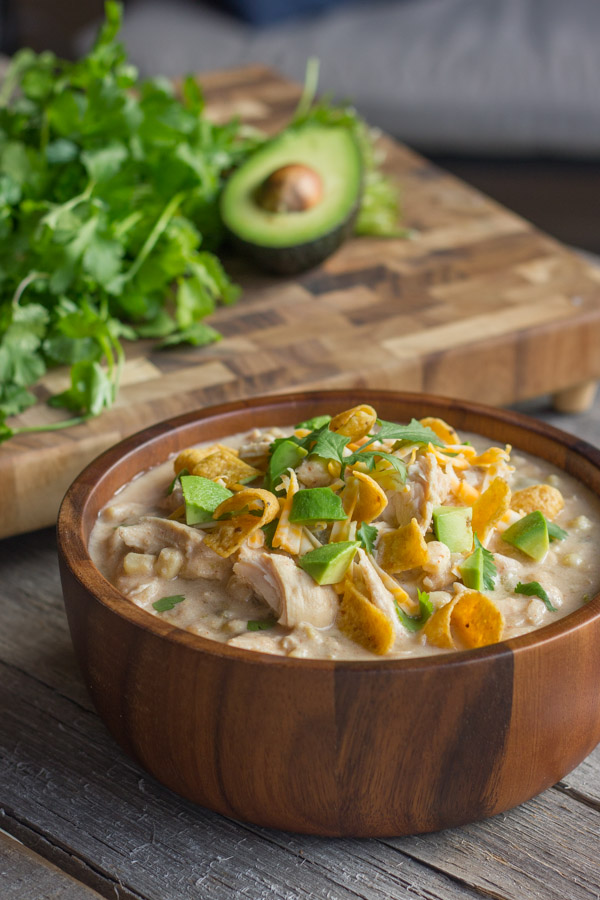 A wooden bowl of Creamy Crockpot White Chicken Chili with fritos, avocado, and cilantro. 