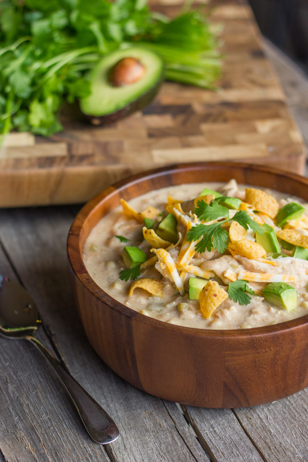 Creamy Crockpot White Chicken Chili in a bowl topped with Fritos, shredded cheese, avocado, and cilantro. There is a spoon next to the bowl, as well as a cutting board with cilantro and half of an avocado in the background. 
