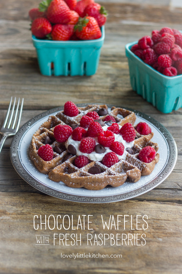 Chocolate Waffles With Fresh Raspberries, topped also with whipped cream and chocolate chips, on a plate with a fork next to it and cartons of fresh berries in the background.