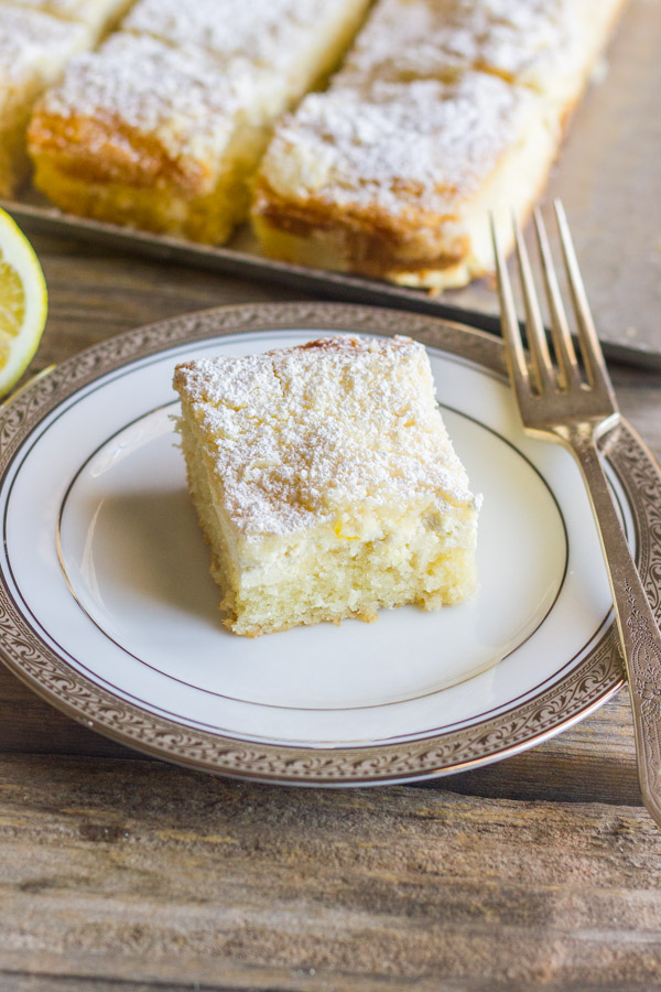 Greek Yogurt Cream Cheese Lemon Coffee Cake square piece on a plate with a fork, and more sliced cake on a serving tray in the background.