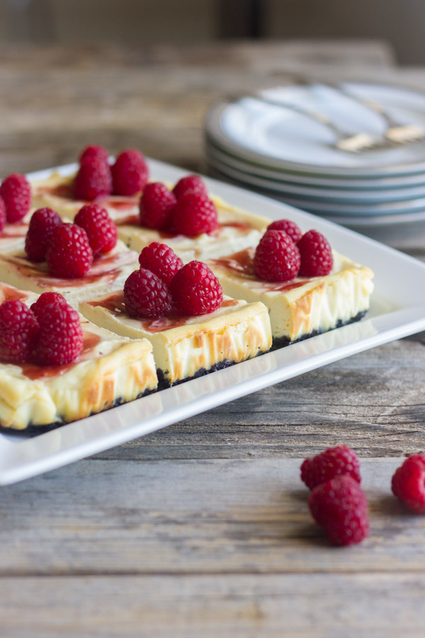 Raspberry Swirl Cheesecake Bars arranged on a serving plate, with a pile of plates in the background with forks on top.