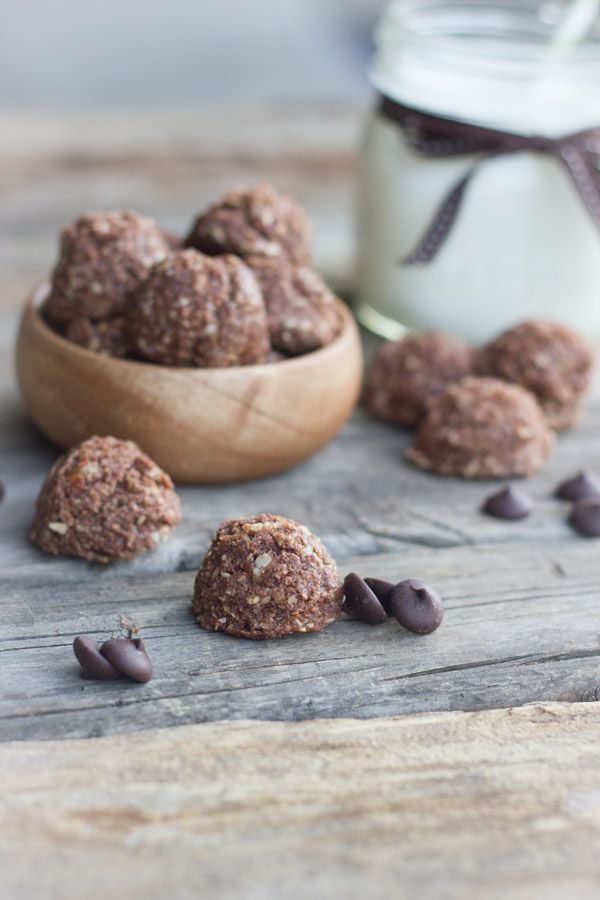 Chocolate Almond Energy Bites piled in a bowl with a few outside of the bowl along with a few chocolate chips and a glass of milk in the background.  