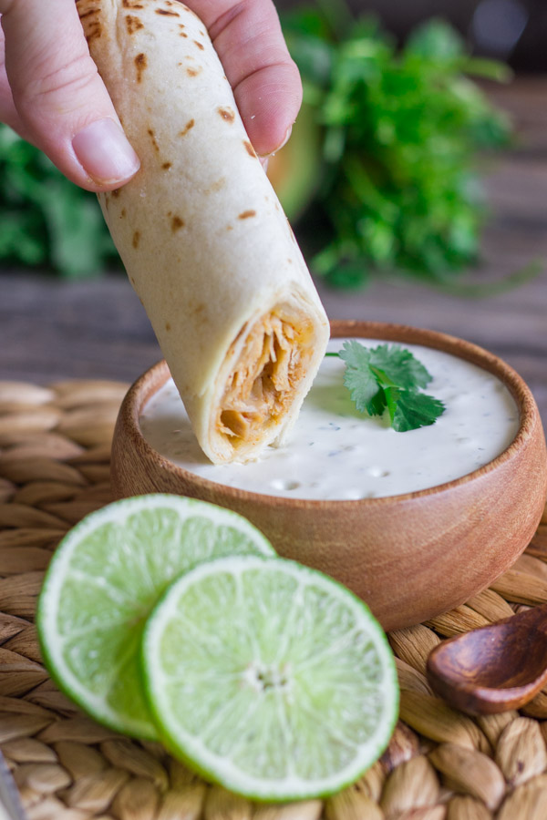 Baked Honey Lime Chicken Taquito being dipped into a small bowl of Cilantro Lime Cream, with a sliced lime next to the bowl.   