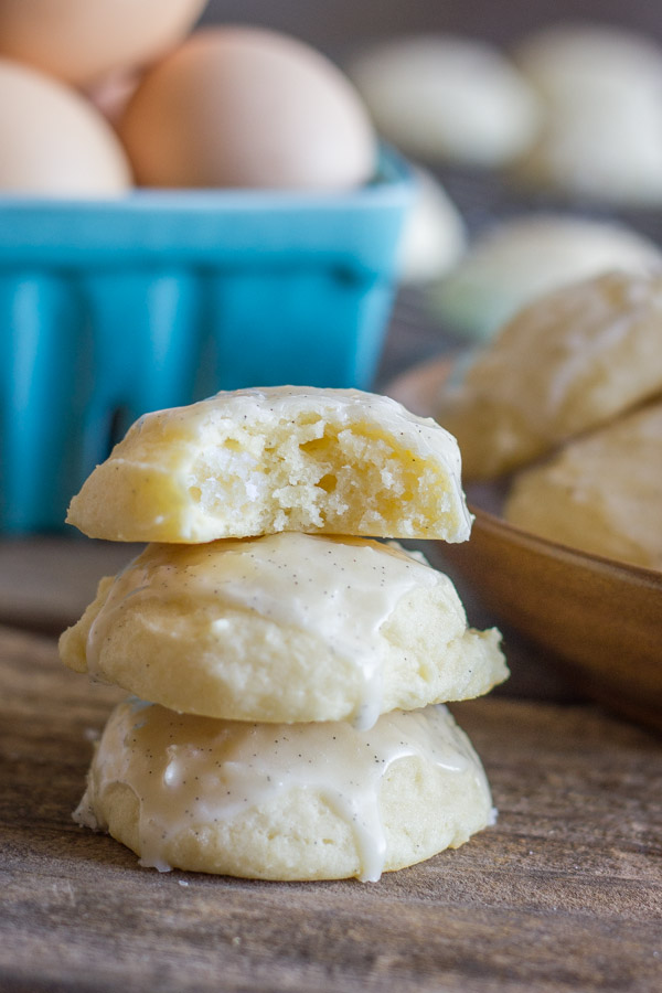 Soft Ricotta Cookies With Vanilla Bean Glaze stacked in a pile of three with a bite taken out of the cookie on top, and a carton of eggs and more cookies in the background.