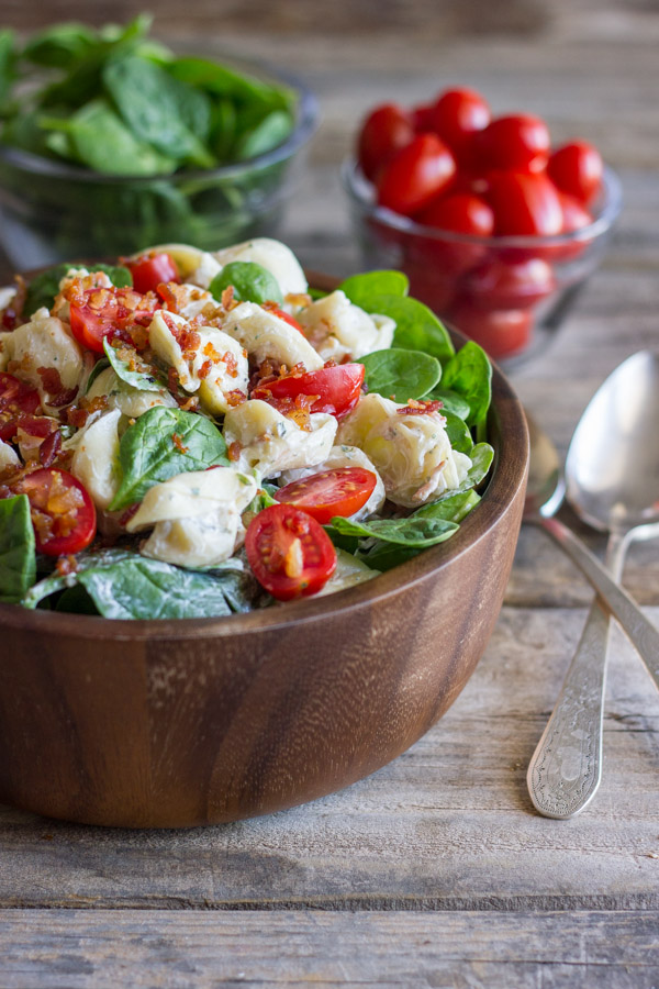 BLT Pasta Salad in a bowl, with a glass dish of spinach, a glass dish of grape tomatoes and two spoons next to the bowl.