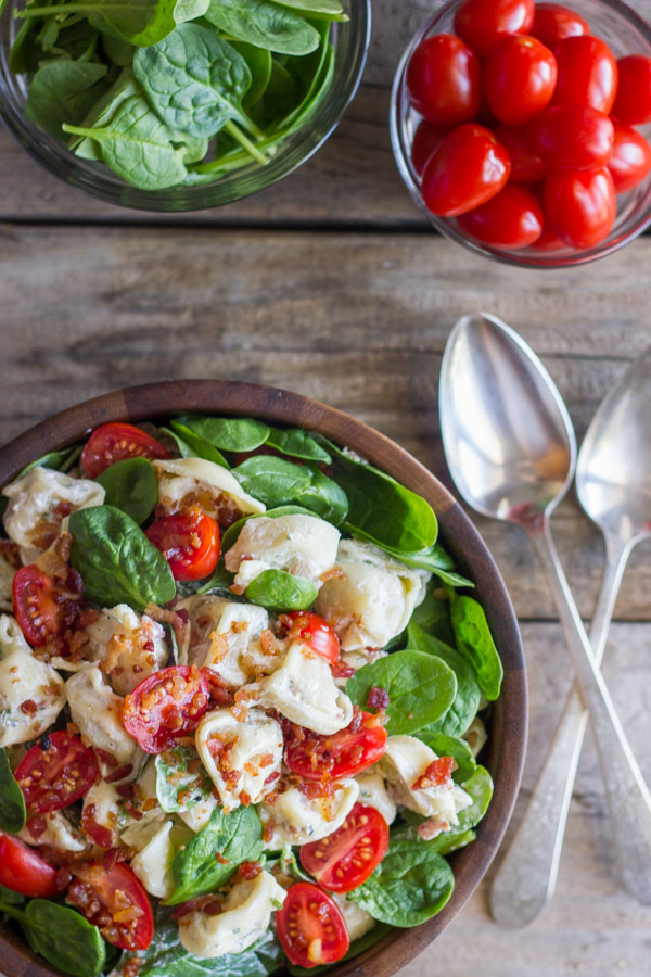 BLT Pasta Salad in a bowl, with a glass dish of spinach, a glass dish of grape tomatoes and two spoons next to the bowl.