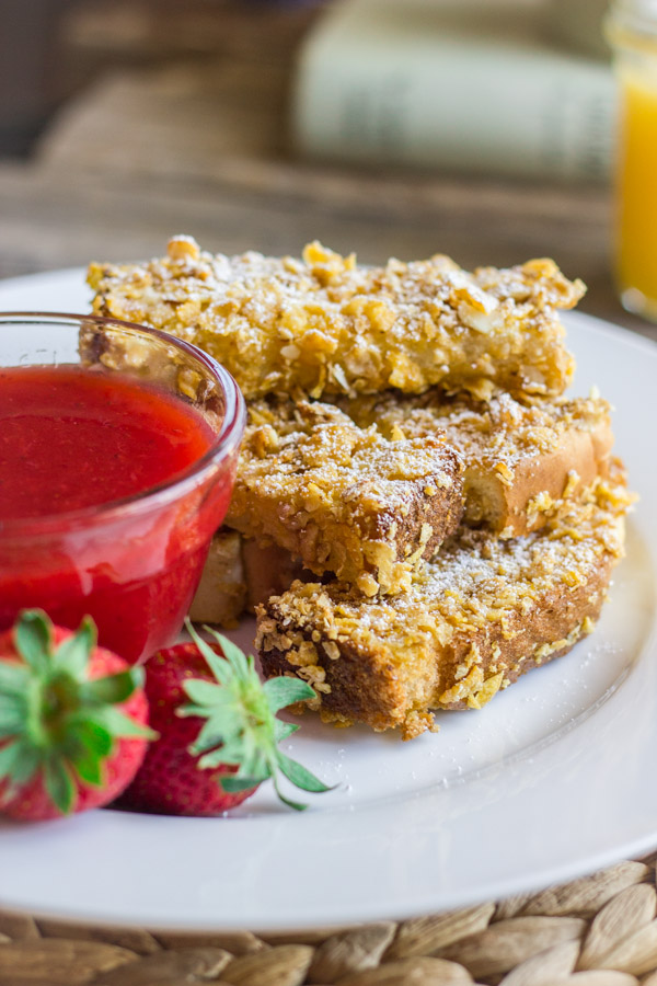 Crunchy Baked French Toast Sticks stacked on a plate next to a glass dish of Strawberry Syrup and some whole strawberries.    