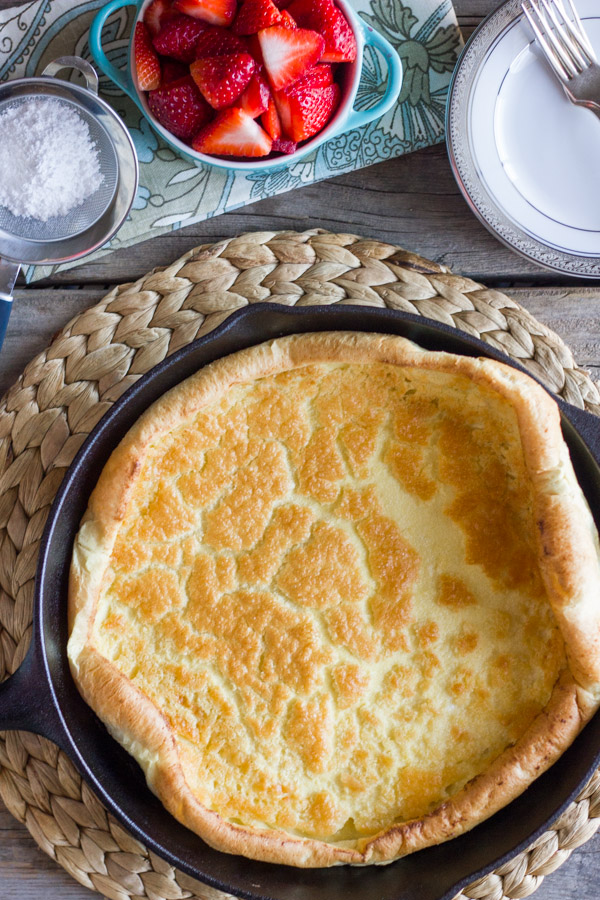 Dutch Baby Pancake in a cast iron skillet, with powdered sugar, a bowl of strawberries and a plate with a fork next to it.  