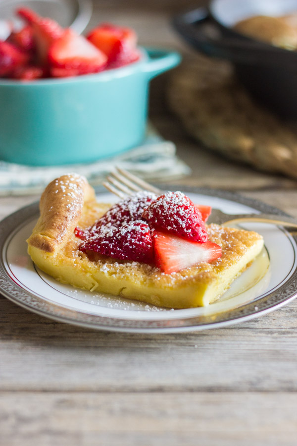 Dutch Baby Pancake slice on a plate topped with strawberries and powdered sugar, with a bowl of strawberries in the background.  