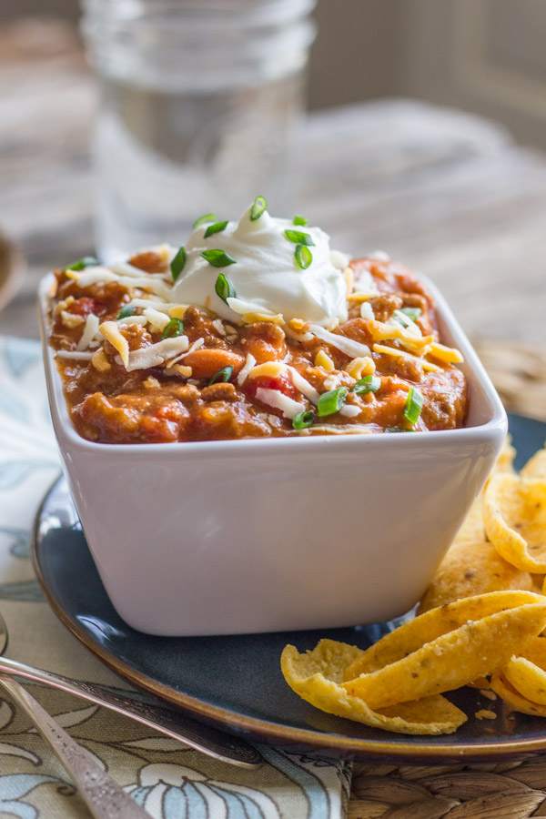 Easy Crockpot Chili in a bowl topped with shredded cheese and sour cream, sitting on a plate with Fritos. 