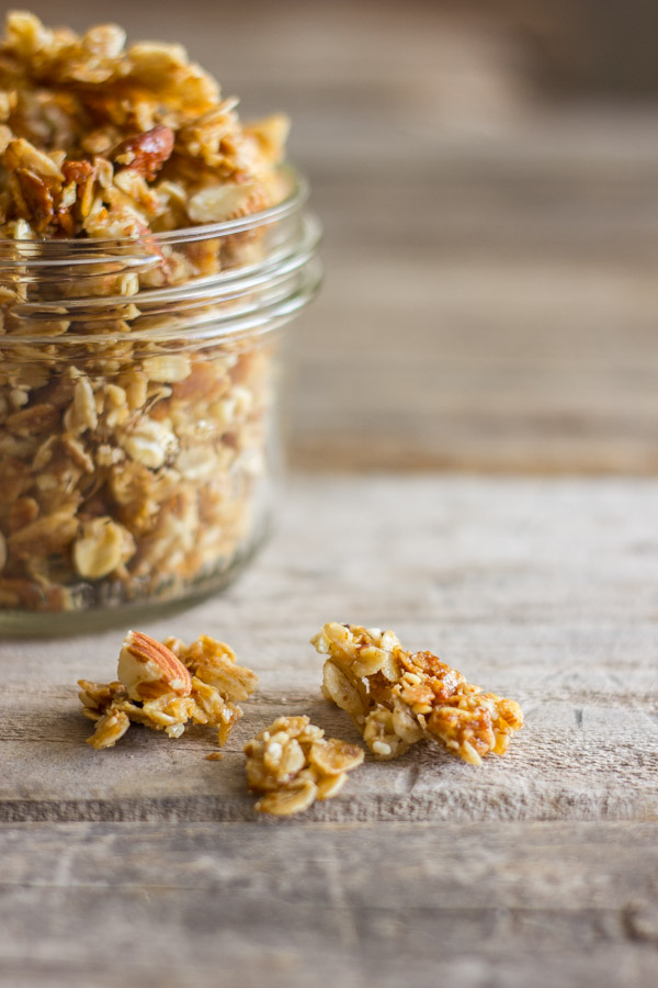 Homemade Coconut Oil Honey Almond Granola in a small glass jar with a few chunks spilled out next to it. 