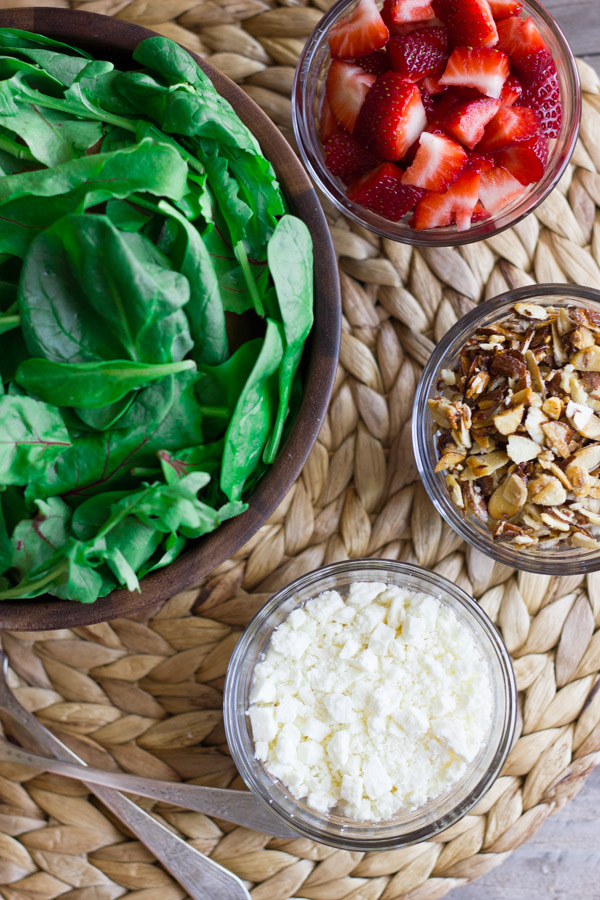 A large bowl of spinach, a small glass dish of strawberries, a small glass dish of sugared almonds, and a small glass dish of Feta cheese.