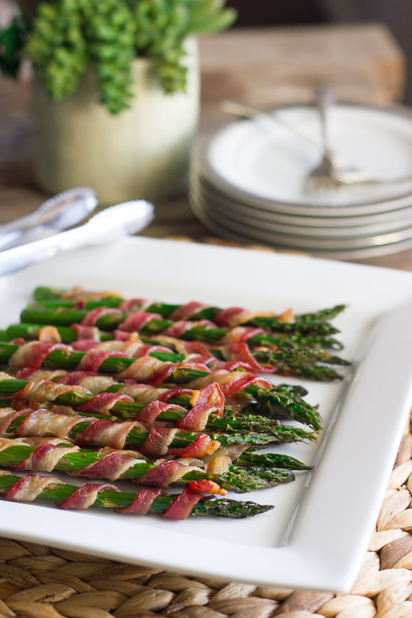 Bacon Wrapped Asparagus on a serving plate, with a stack of plates and forks in the background.  