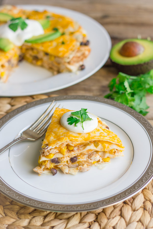 Southwestern Chicken Taco Pie slice topped with sour cream and cilantro, on a plate with a fork.  A plate of Southwestern Chicken Taco Pie topped with sour cream, avocado slices and cilantro in the background, along with a half of an avocado and some cilantro.  
