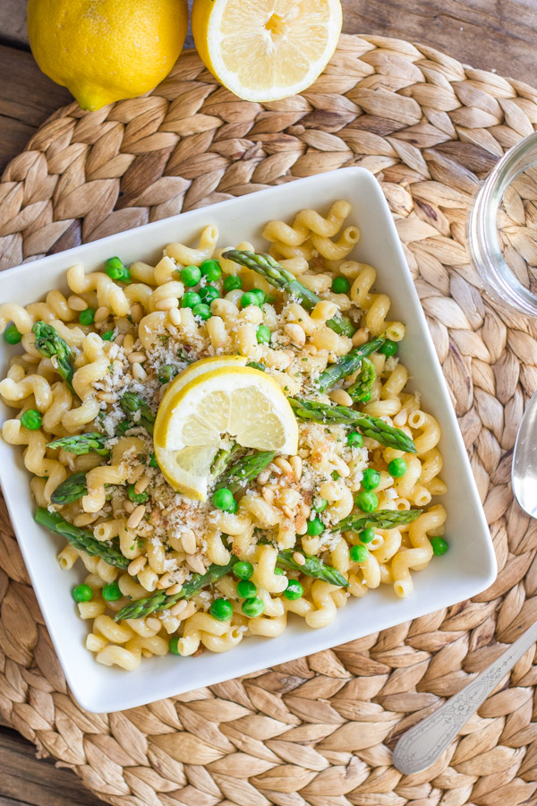 Springtime Pasta With Brown Butter Garlic Cream Sauce in a square bowl, topped with homemade breadcrumbs and a lemon wedge, with a spoon and water glass next to it, along with some lemons.