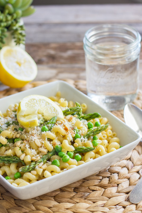 Springtime Pasta With Brown Butter Garlic Cream Sauce in a square bowl, topped with homemade breadcrumbs and a lemon wedge, with a spoon and water glass next to it, and a half of a lemon in the background.