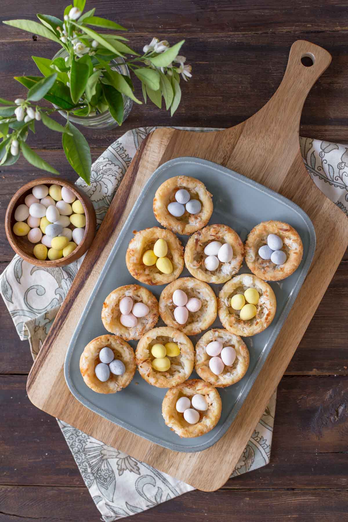 Sugar Cookie Easter Egg Nests on a serving plate, with a bowl of mini Cadbury chocolate eggs next to it along with a vase of greenery.  