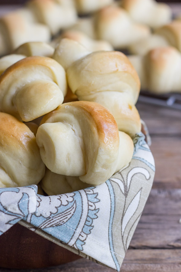 The Best Butterhorn Rolls in a cloth lined wooden bowl, with more Butterhorn Rolls on a cooling rack in the background.  