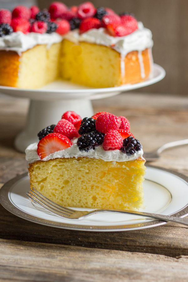A slice of Easy Triple Berry Almond Poke Cake on a plate with a fork, and the rest of the cake on a cake stand in the background.  