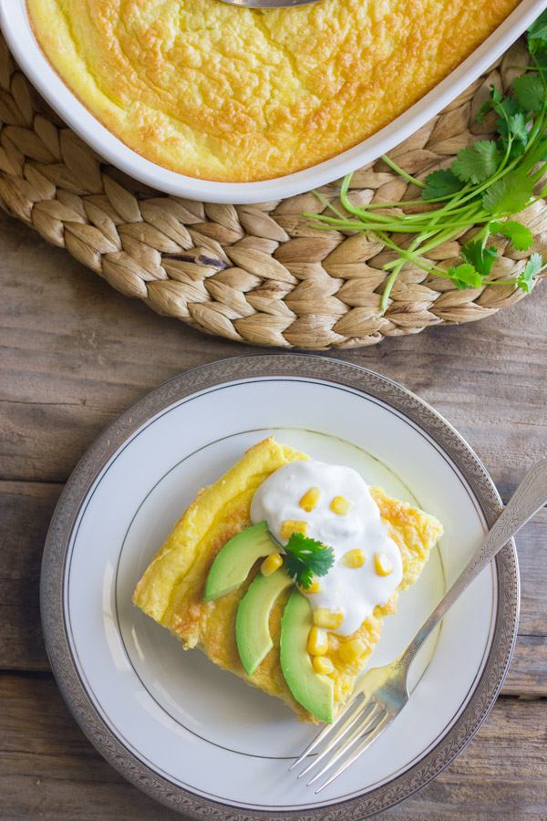 Green Chile Egg Bake Made With Greek Yogurt topped with avocado slices, sour cream, corn kernels and cilantro, on a plate with a fork, sitting next to the baking dish of the egg bake and a bundle of cilantro. 