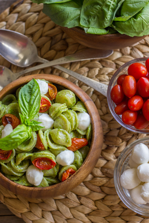 Orecchiette With Pesto and Oven Roasted Tomatoes in a bowl, topped with fresh basil leaves, sitting next to a bowl of fresh mozzarella, a bowl of grape tomatoes, a bowl of fresh basil leaves and two spoons.