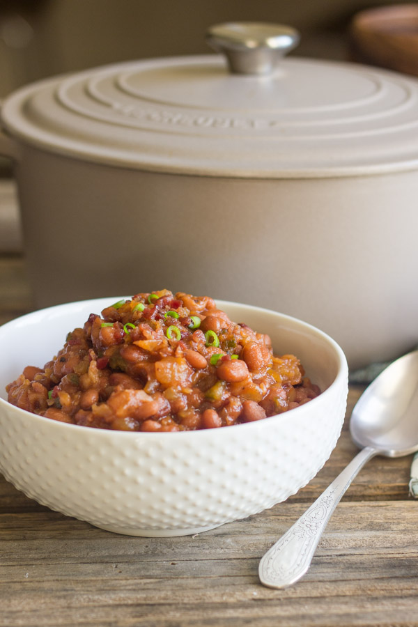 Sweet And Spicy BBQ Baked Beans in a bowl with a spoon next to it and a Le Creuset French Oven in the background.