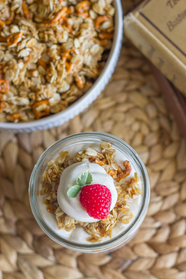 Raspberry Pretzel Granola Parfait in a glass jar, sitting next to a bowl of Pretzel Granola and a stack of books.