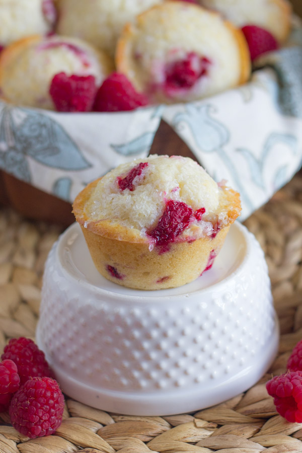 Healthier Raspberry Almond Muffin sitting on top of a glass dish with fresh raspberries around it, and a cloth lined bowl full of muffins in the background.