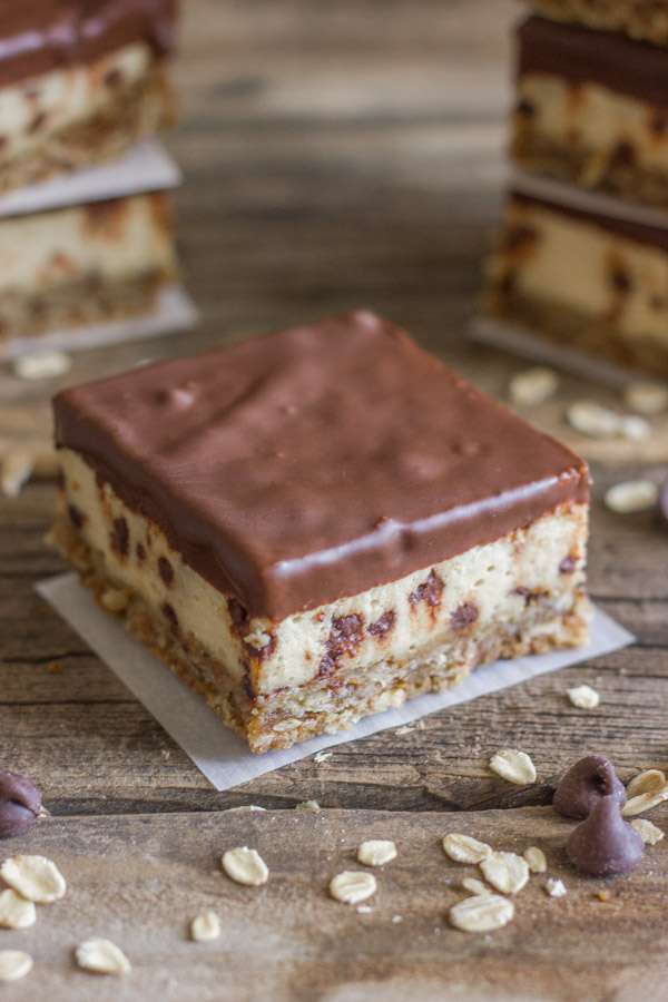 Oatmeal Chocolate Chip Cheesecake Bar on a board with oats and chocolate chips on it, with more Oatmeal Chocolate Chip Cheesecake Bars stacked in the background.