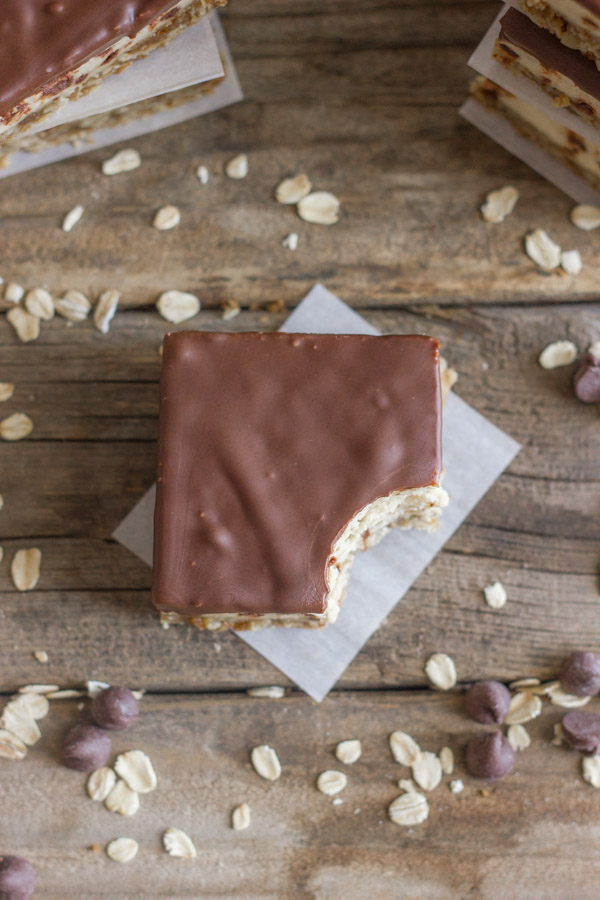 Oatmeal Chocolate Chip Cheesecake Bar with a bite out of it on a board with oats and chocolate chips on it, with more Oatmeal Chocolate Chip Cheesecake Bars stacked next to it.
