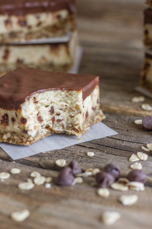 Oatmeal Chocolate Chip Cheesecake Bar with a bite out of it on a board with oats and chocolate chips on it, with more Oatmeal Chocolate Chip Cheesecake Bars stacked in the background.