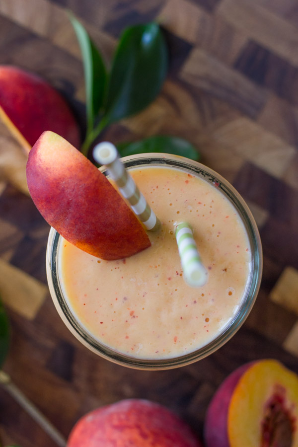 Skinny Peach Cream Slush in a glass with two straws and a peach slice, sitting on a cutting board with peaches around it.  
