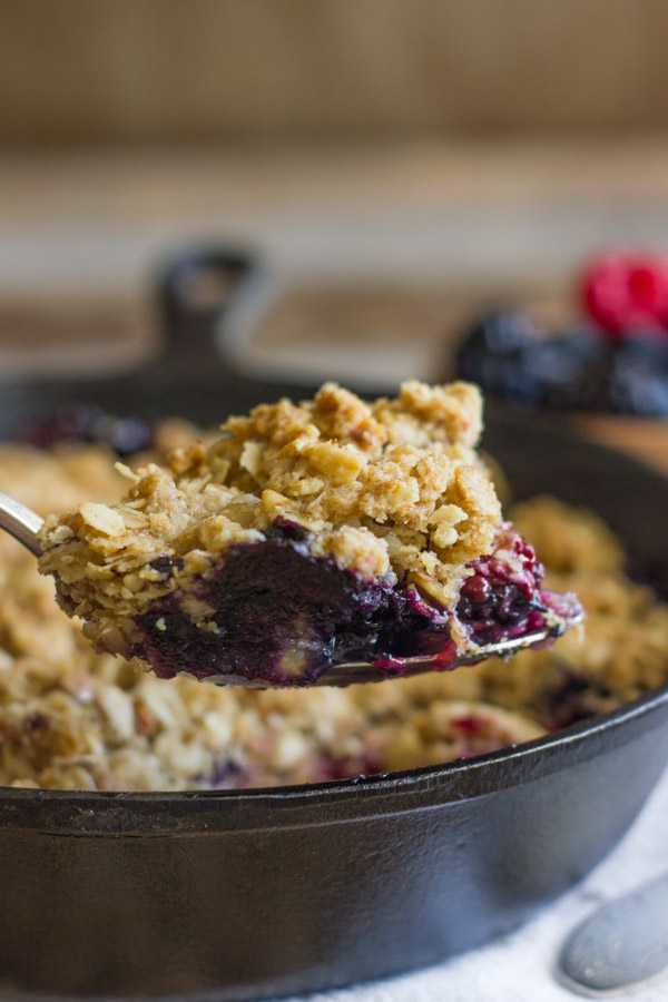 Triple Berry Skillet Crisp on a serving spoon, with the cast iron skillet of Triple Berry Skillet Crisp in the background.  