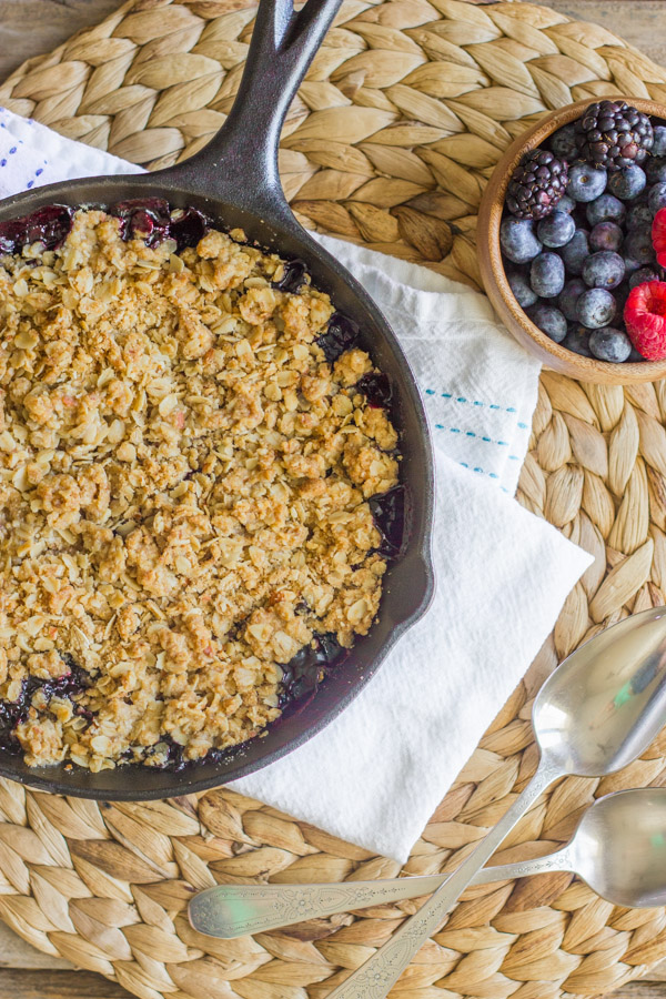 Triple Berry Skillet Crisp in a cast iron skillet, with a bowl of berries and spoons next to it.