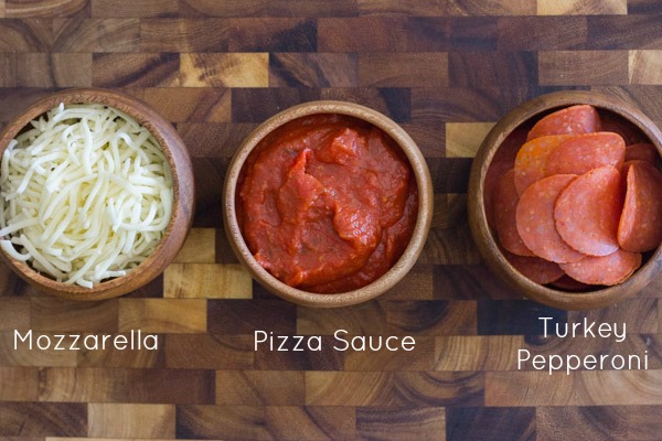 A bowl of mozzarella cheese, a bowl of pizza sauce, and a bowl of turkey pepperoni all sitting on a cutting board.