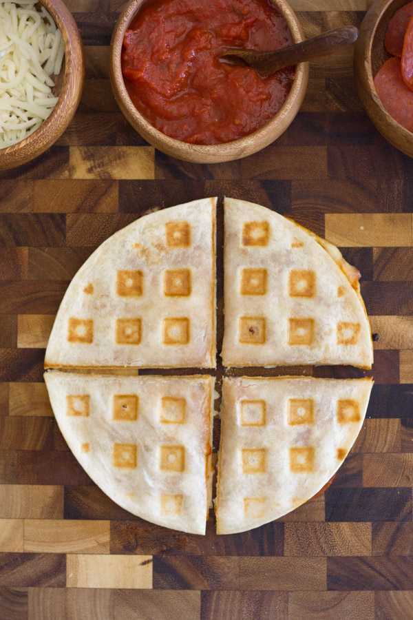 Waffled Pizza Pocket cut in four pieces on at cutting board with a bowl of mozzarella cheese, a bowl of pizza sauce and a bowl of pepperoni next to it.