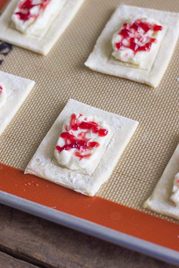 Puff pastry dough on a Silpat lined baking sheet, with sweetened cream cheese mixture placed onto the center of the pastry dough along with a swirl of raspberry jam.