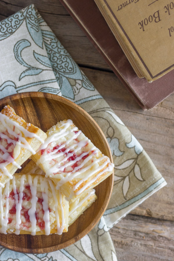 Raspberry Cream Cheese Danishes on a wood plate that is sitting on a cloth napkin, with some stacked books sitting next to them.  