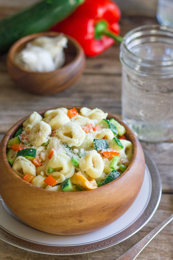 Vegetable Tortellini With Creamy Garlic Sauce in a wood bowl sitting on a plate, with a glass of water, a small wood bowl of garlic bulbs, a zucchini and a red pepper in the background.