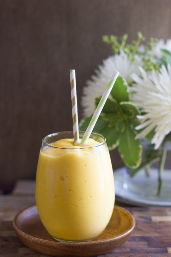 Tropical Sunshine Smoothie in a glass with two straws sitting on a little wood saucer, with a flower arrangement in the background.  