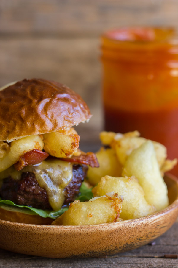 Western Bacon Sliders With Fried Cheese Curds on a wood plate, with a glass jar of BBQ sauce in the background.  