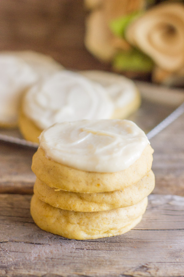 A stack of three Soft Pumpkin Cookies With Maple Cream Cheese Frosting, with more cookies on a platter in the background.  