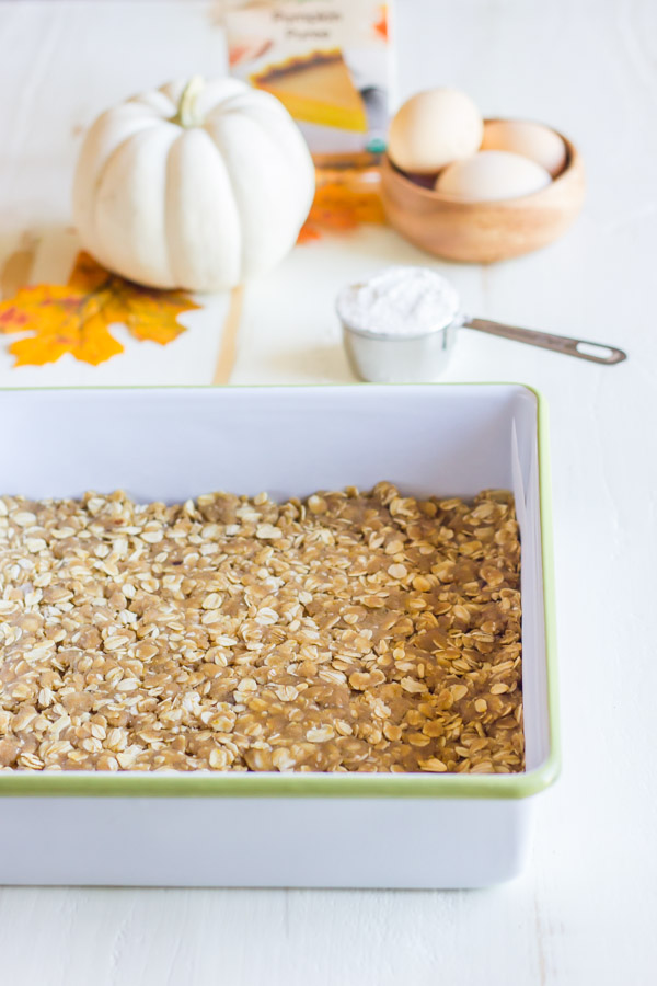 Oatmeal cookie crust for the Pumpkin Pie Cheesecake Bars in a baking dish, with more ingredients in the background.  
