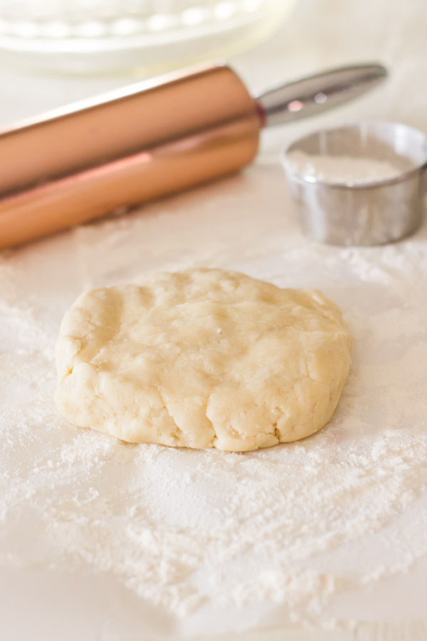 Pie Crust Tutorial Step 4 - pie dough on top of floured parchment paper, with a rolling pin and a cup of flour in the background.