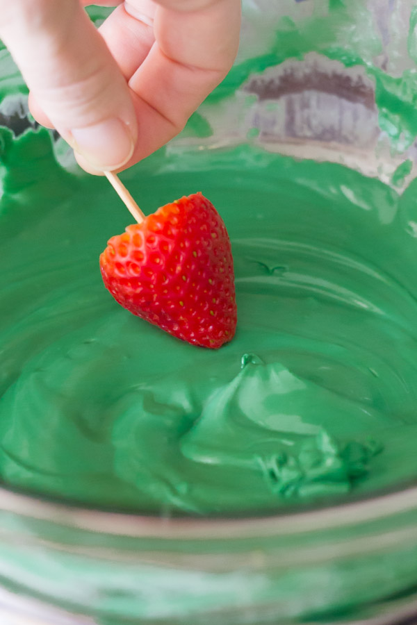 A strawberry on a toothpick being dipped into the melted candy melts. 