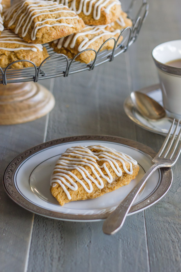 Iced Gingerbread Oat Scone on a plate with a fork, and more scones on a cake stand in the background along with a cup of coffee.  